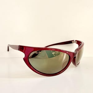 May include: Maroon-colored Ray-Ban sunglasses with a unique, curved frame design. The lenses are a dark, reflective color. The Ray-Ban logo is visible on the left lens. The sunglasses are set against a white background.
