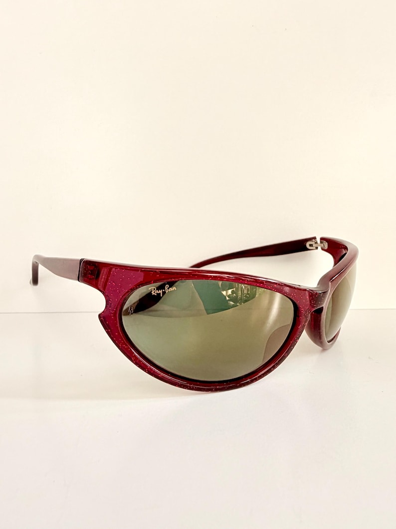 May include: Maroon-colored Ray-Ban sunglasses with a unique, curved frame design. The lenses are a dark, reflective color. The Ray-Ban logo is visible on the left lens. The sunglasses are set against a white background.