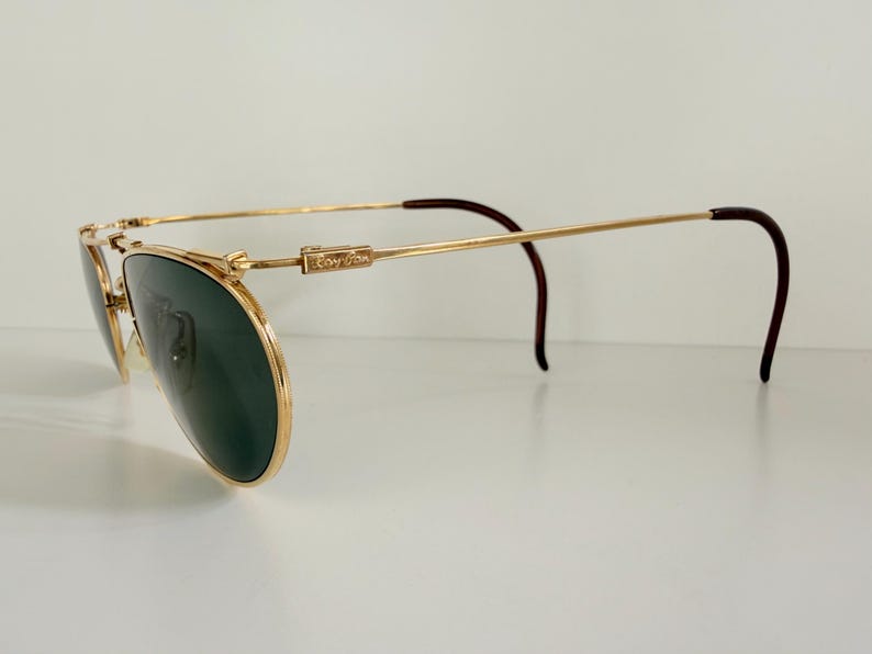 K&ouml;nnte beinhalten: Goldfarbene Ray-Ban Aviator-Sonnenbrille mit dunkelgr&uuml;nen Gl&auml;sern. Die Fassungen haben ein klassisches Doppelsteg-Design und braune B&uuml;gelenden. Das Ray-Ban-Logo ist auf dem B&uuml;gel zu sehen. Ein stilvolles Accessoire zum Schutz der Augen.