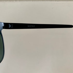 May include: Close-up of black sunglasses with green lenses. The frame is black, and the text "W0757" is printed on the temple arm. The sunglasses are set against a neutral background.