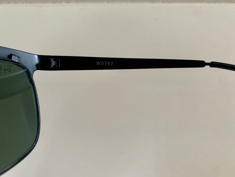May include: Close-up of black sunglasses with green lenses. The frame is black, and the text "W0757" is printed on the temple arm. The sunglasses are set against a neutral background.