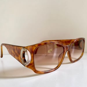 May include: Brown tortoiseshell sunglasses with gradient lenses. The sunglasses have a large, square frame with a silver accent on the temples. The text "Christian Dior" is visible on the silver accent.