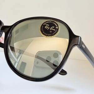 May include: Black Ray-Ban sunglasses with round frames. The lenses are a light green color. The Ray-Ban logo is visible on the lens. The sunglasses have a classic, timeless design.