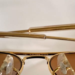 May include: Close-up of gold-coloured Ray-Ban sunglasses with brown lenses. The text "B&L RAY-BAN 62[]14" is visible on the frame. The sunglasses have a classic aviator style with a double bridge and clear nose pads.