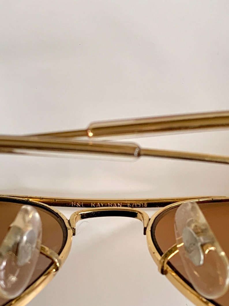 May include: Close-up of gold-coloured Ray-Ban sunglasses with brown lenses. The text "B&L RAY-BAN 62[]14" is visible on the frame. The sunglasses have a classic aviator style with a double bridge and clear nose pads.