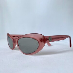 Peut inclure: Lunettes de soleil roses en forme d'&oelig;il de chat avec des verres gris. Les lunettes ont une monture translucide et un petit logo sur les branches. Les verres sont gris fonc&eacute; et la monture est rose clair.