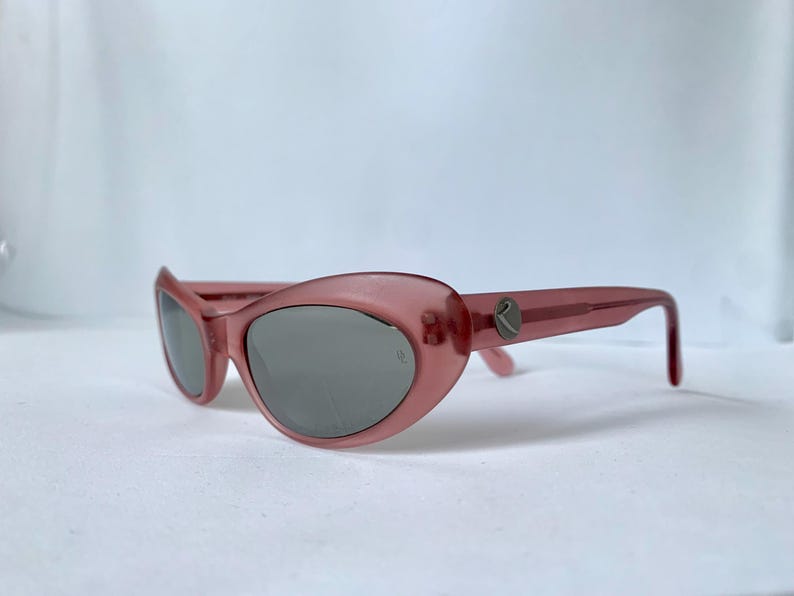 Peut inclure: Lunettes de soleil roses en forme d'&oelig;il de chat avec des verres gris. Les lunettes ont une monture translucide et un petit logo sur les branches. Les verres sont gris fonc&eacute; et la monture est rose clair.