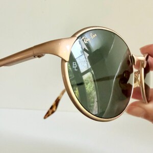 Puede incluir: Gafas de sol Ray-Ban doradas con lentes redondas de color verde oscuro. Las monturas son de color oro claro y las patillas tienen un dise&ntilde;o de carey. El logotipo de Ray-Ban es visible en la lente.
