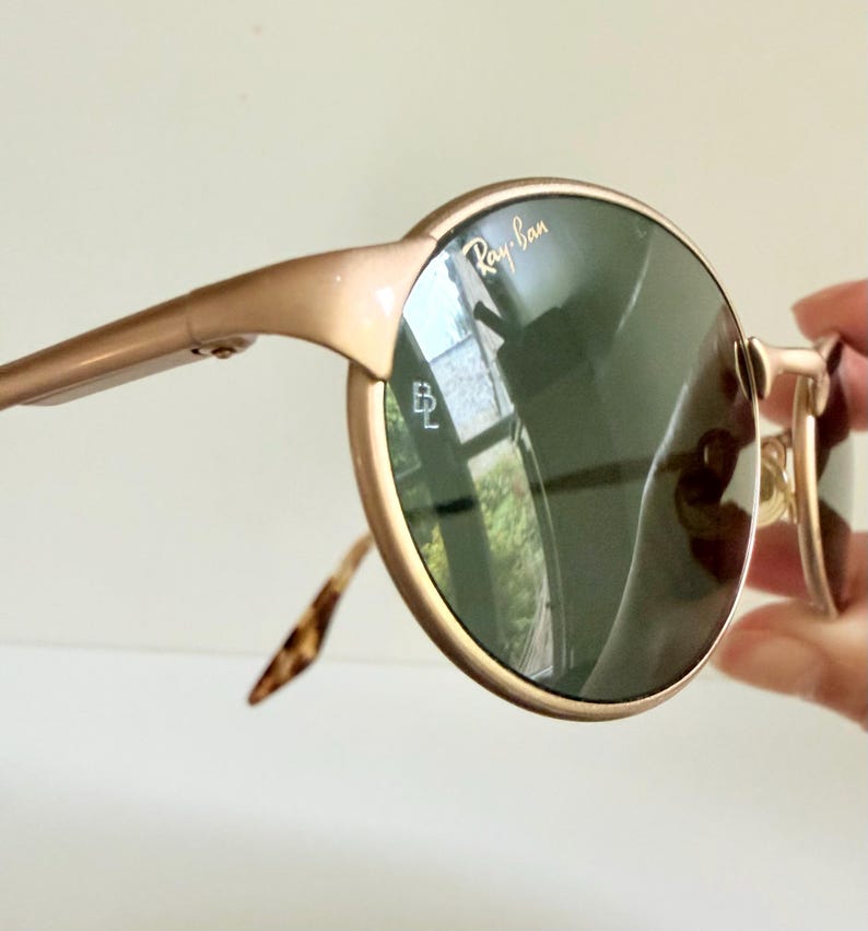 Puede incluir: Gafas de sol Ray-Ban doradas con lentes redondas de color verde oscuro. Las monturas son de color oro claro y las patillas tienen un dise&ntilde;o de carey. El logotipo de Ray-Ban es visible en la lente.