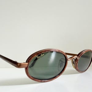 May include: Oval sunglasses with a brown marbled frame and dark green lenses. The Ray-Ban logo is visible on the lens. The temples are a dark brown colour. The sunglasses are resting on a white surface.