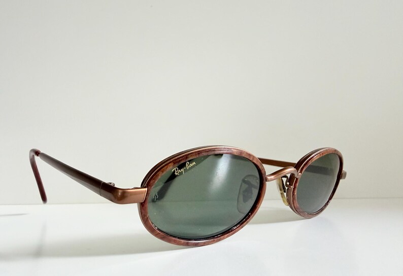 May include: Oval sunglasses with a brown marbled frame and dark green lenses. The Ray-Ban logo is visible on the lens. The temples are a dark brown colour. The sunglasses are resting on a white surface.