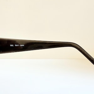 May include: Close-up of a black Ray-Ban sunglasses arm. The arm is glossy and features the text "B&L RAY-BAN" in white. The sunglasses have a classic design, suitable for various styles.