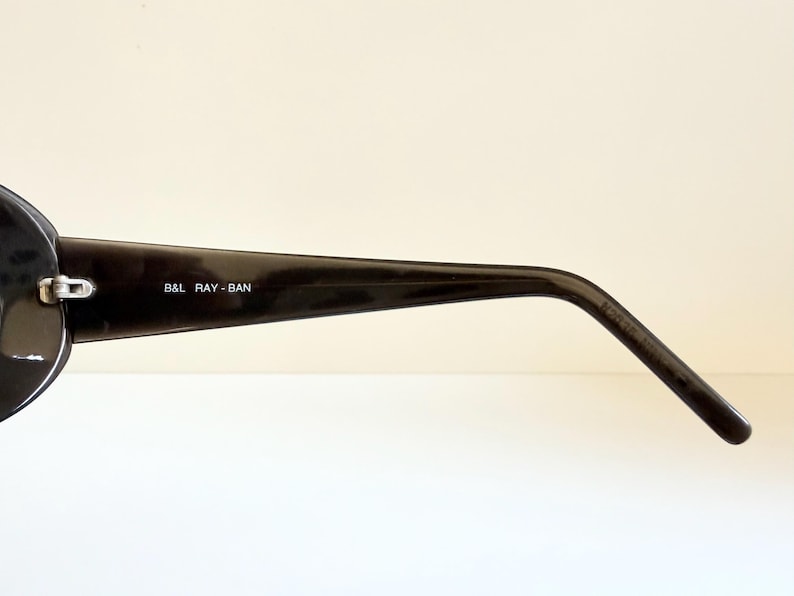 May include: Close-up of a black Ray-Ban sunglasses arm. The arm is glossy and features the text "B&L RAY-BAN" in white. The sunglasses have a classic design, suitable for various styles.