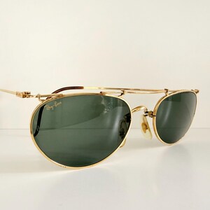 K&ouml;nnte beinhalten: Goldfarbene Sonnenbrille mit gr&uuml;nen Gl&auml;sern. Die Sonnenbrille hat einen Doppelsteg und ein klassisches Design. Das Ray-Ban-Logo ist auf dem Glas sichtbar. Die B&uuml;gel sind dunkelbraun.