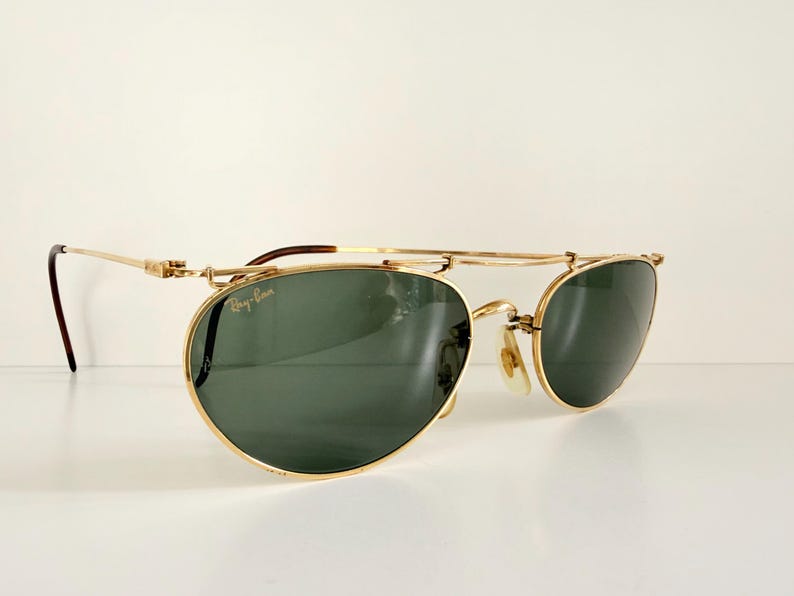 K&ouml;nnte beinhalten: Goldfarbene Sonnenbrille mit gr&uuml;nen Gl&auml;sern. Die Sonnenbrille hat einen Doppelsteg und ein klassisches Design. Das Ray-Ban-Logo ist auf dem Glas sichtbar. Die B&uuml;gel sind dunkelbraun.