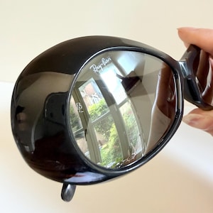 May include: Black Ray-Ban sunglasses with a distinctive, rounded frame. The lenses are reflective, displaying a mirrored image. The Ray-Ban logo is visible on the lens. The sunglasses are held by a person's hand.
