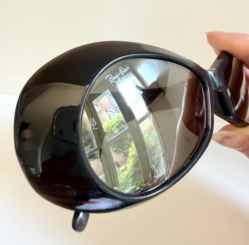 May include: Black Ray-Ban sunglasses with a distinctive, rounded frame. The lenses are reflective, displaying a mirrored image. The Ray-Ban logo is visible on the lens. The sunglasses are held by a person's hand.