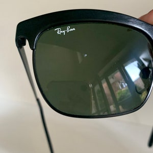 May include: Black sunglasses with green lenses. The Ray-Ban logo is visible in white on the upper left of the frame. The sunglasses have a classic design with a thick, dark frame and thin arms.