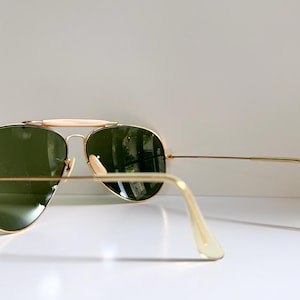 May include: Gold-framed aviator sunglasses with green lenses. The sunglasses have a double bridge and a light-colored brow bar. The temples are thin and gold, with light-colored ear pieces. The lenses reflect the surrounding environment.