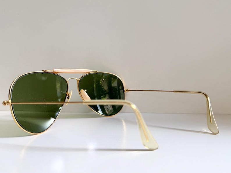 May include: Gold-framed aviator sunglasses with green lenses. The sunglasses have a double bridge and a light-colored brow bar. The temples are thin and gold, with light-colored ear pieces. The lenses reflect the surrounding environment.