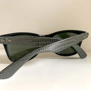 May include: Black sunglasses with gray, textured arms. The sunglasses have dark lenses and the Ray-Ban logo is visible on the arm. The arms have a reptile-like pattern. The sunglasses are angled, showing the inside of the lenses.