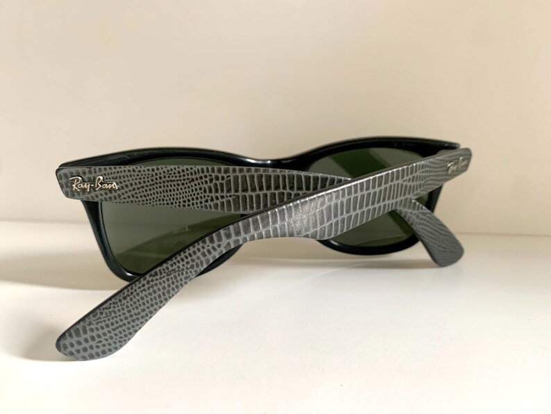 May include: Black sunglasses with gray, textured arms. The sunglasses have dark lenses and the Ray-Ban logo is visible on the arm. The arms have a reptile-like pattern. The sunglasses are angled, showing the inside of the lenses.
