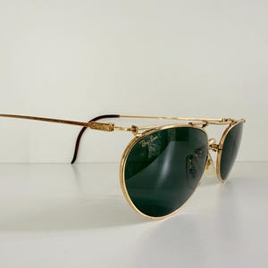 K&ouml;nnte beinhalten: Goldfarbene Sonnenbrille mit dunkelgr&uuml;nen Gl&auml;sern. Die Sonnenbrille hat einen Doppelsteg und gebogene B&uuml;gel mit braunen Spitzen. Das Ray-Ban-Logo ist auf dem Glas zu sehen. Die Brille steht vor wei&szlig;em Hintergrund.