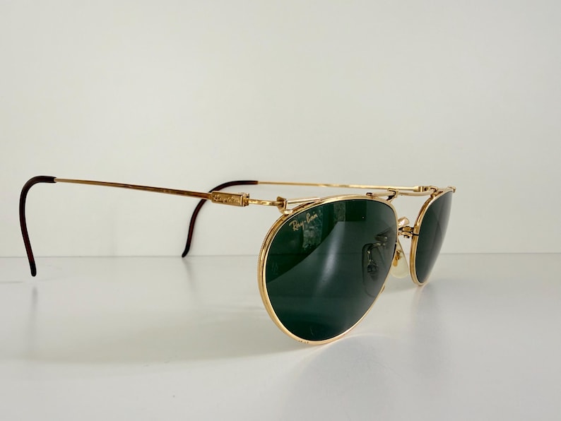 K&ouml;nnte beinhalten: Goldfarbene Sonnenbrille mit dunkelgr&uuml;nen Gl&auml;sern. Die Sonnenbrille hat einen Doppelsteg und gebogene B&uuml;gel mit braunen Spitzen. Das Ray-Ban-Logo ist auf dem Glas zu sehen. Die Brille steht vor wei&szlig;em Hintergrund.