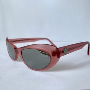 Peut inclure: Lunettes de soleil roses avec une forme arrondie en &oelig;il de chat et des verres gris. Les montures ont une qualit&eacute; subtile et translucide. Un petit logo argent&eacute; est visible sur les branches. Les lunettes sont sur fond blanc.