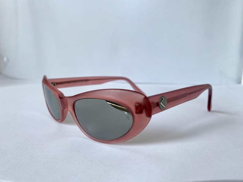 Peut inclure: Lunettes de soleil roses avec une forme arrondie en &oelig;il de chat et des verres gris. Les montures ont une qualit&eacute; subtile et translucide. Un petit logo argent&eacute; est visible sur les branches. Les lunettes sont sur fond blanc.