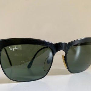 May include: Black sunglasses with green lenses. The Ray-Ban logo is visible on the upper left lens. The sunglasses have a classic design with a textured black frame.