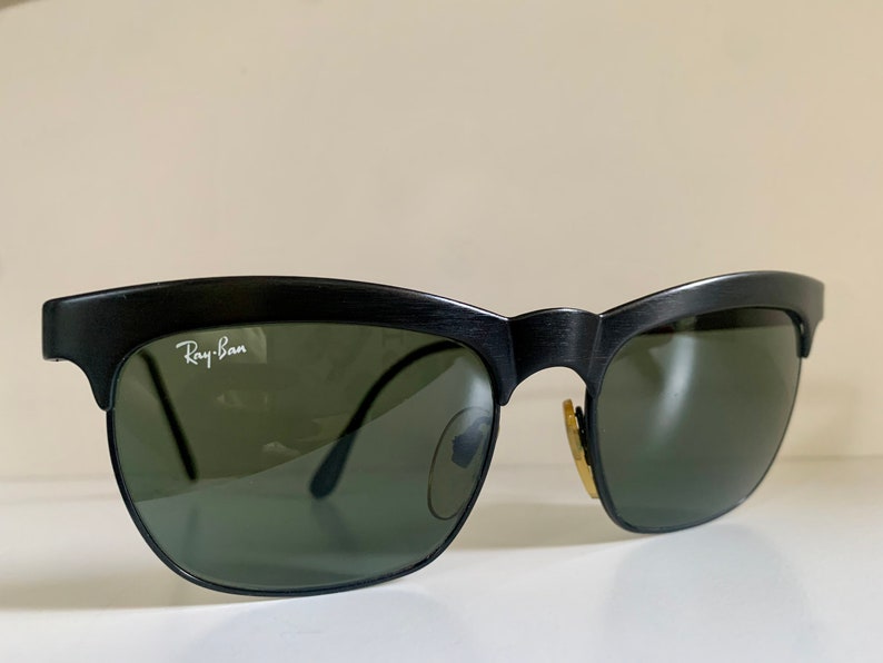 May include: Black sunglasses with green lenses. The Ray-Ban logo is visible on the upper left lens. The sunglasses have a classic design with a textured black frame.