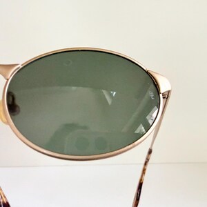 Puede incluir: Gafas de sol ovaladas con montura dorada y lentes verdes. Las patillas tienen un dise&ntilde;o marr&oacute;n y beige. Estas gafas de sol est&aacute;n dise&ntilde;adas para proteger los ojos del sol.