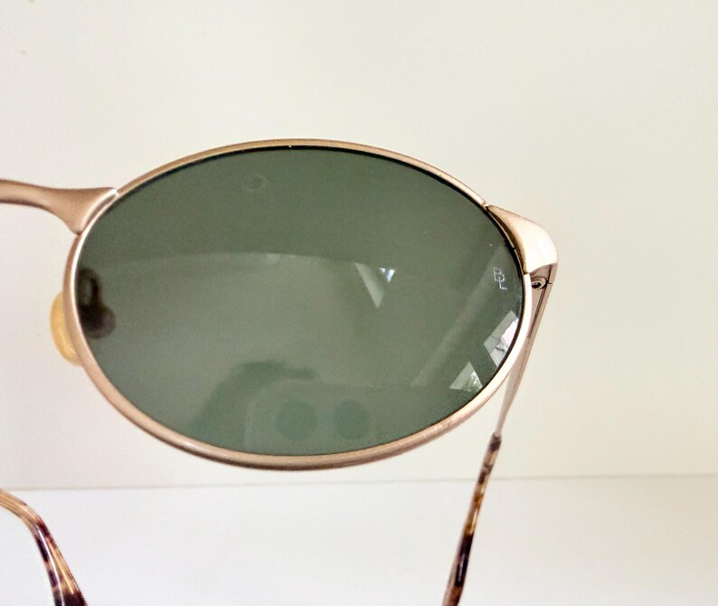Puede incluir: Gafas de sol ovaladas con montura dorada y lentes verdes. Las patillas tienen un dise&ntilde;o marr&oacute;n y beige. Estas gafas de sol est&aacute;n dise&ntilde;adas para proteger los ojos del sol.