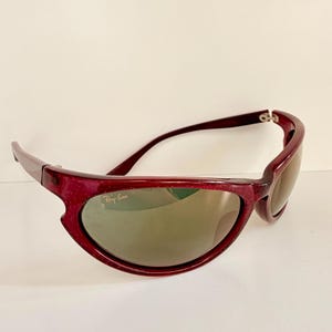 May include: Maroon-colored Ray-Ban sunglasses with a curved frame and tinted lenses. The sunglasses have a sporty design and the Ray-Ban logo is visible on the lens.