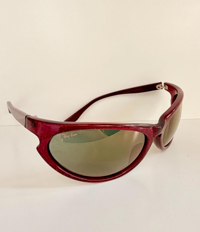 May include: Maroon-colored Ray-Ban sunglasses with a curved frame and tinted lenses. The sunglasses have a sporty design and the Ray-Ban logo is visible on the lens.