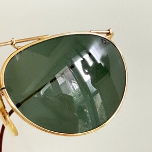 K&ouml;nnte beinhalten: Nahaufnahme einer Vintage-Sonnenbrille mit goldfarbenem Metallrahmen und dunkelgr&uuml;nen Gl&auml;sern. Die Gl&auml;ser haben eine leichte Kr&uuml;mmung und reflektieren die Umgebung. Die Sonnenbrille hat einen klassischen Aviator-Stil.