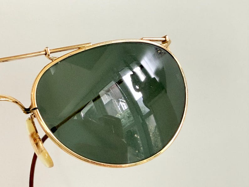 K&ouml;nnte beinhalten: Nahaufnahme einer Vintage-Sonnenbrille mit goldfarbenem Metallrahmen und dunkelgr&uuml;nen Gl&auml;sern. Die Gl&auml;ser haben eine leichte Kr&uuml;mmung und reflektieren die Umgebung. Die Sonnenbrille hat einen klassischen Aviator-Stil.