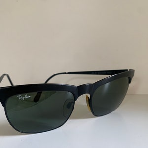 May include: Black Ray-Ban sunglasses with a semi-rimless design. The sunglasses have a matte black frame, green lenses, and the Ray-Ban logo in white on the left lens. The temples are thin and black. The sunglasses are set against a neutral background.