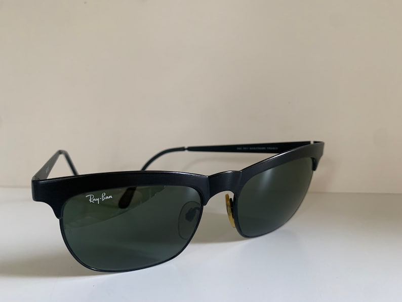 May include: Black Ray-Ban sunglasses with a semi-rimless design. The sunglasses have a matte black frame, green lenses, and the Ray-Ban logo in white on the left lens. The temples are thin and black. The sunglasses are set against a neutral background.