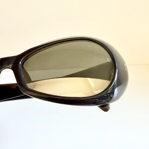 May include: Black sunglasses with a rounded frame and tinted lenses. The sunglasses have a glossy finish and a small detail on the side of the frame. The lenses are a gradient colour.