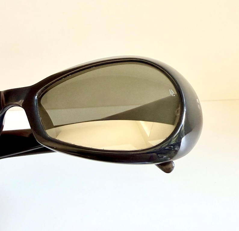 May include: Black sunglasses with a rounded frame and tinted lenses. The sunglasses have a glossy finish and a small detail on the side of the frame. The lenses are a gradient colour.