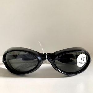 May include: Black sunglasses with oval lenses. The sunglasses have a thick, curved frame and a small white sticker with text. The sunglasses are on a white surface.