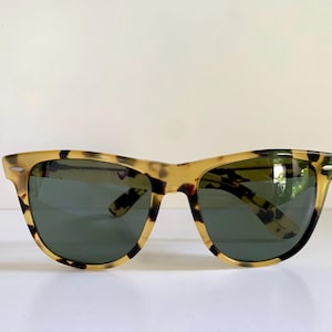 May include: Tortoiseshell sunglasses with dark green lenses. The frames have a classic, angular shape with a speckled pattern in shades of brown and tan. The sunglasses are set against a white background.