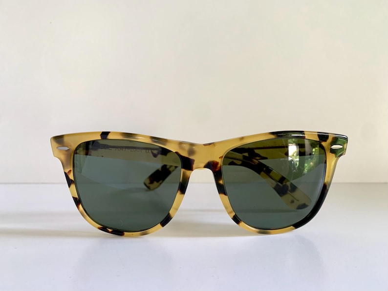 May include: Tortoiseshell sunglasses with dark green lenses. The frames have a classic, angular shape with a speckled pattern in shades of brown and tan. The sunglasses are set against a white background.