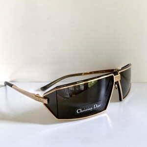 Peut inclure: Lunettes de soleil &agrave; monture dor&eacute;e avec verres fonc&eacute;s. Les lunettes ont une forme rectangulaire avec une monture dor&eacute;e et le texte "Christian Dior" sur le verre. Les branches sont dor&eacute;es avec des embouts noirs.