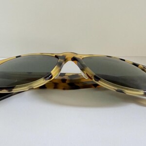 May include: Sunglasses with a classic design. The frames have a tortoiseshell pattern in shades of tan and brown. The lenses are dark, providing shade and protection from the sun. The arms are extended.