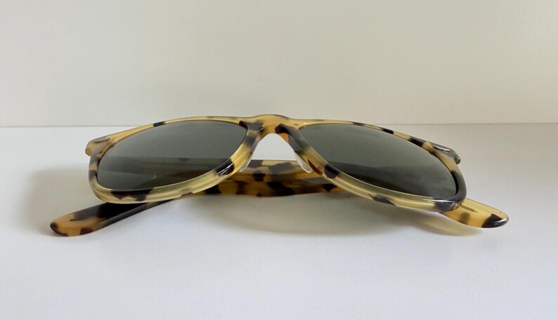 May include: Sunglasses with a classic design. The frames have a tortoiseshell pattern in shades of tan and brown. The lenses are dark, providing shade and protection from the sun. The arms are extended.