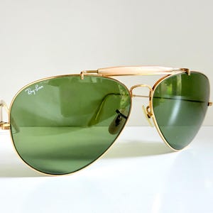 May include: Gold-framed aviator sunglasses with green lenses. The Ray-Ban logo is visible on the left lens. The sunglasses have a double bridge and a light-colored bar above the nose bridge.
