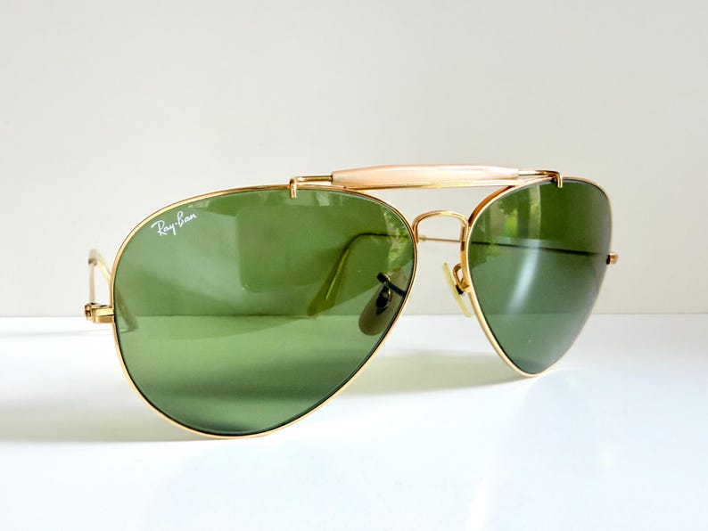 May include: Gold-framed aviator sunglasses with green lenses. The Ray-Ban logo is visible on the left lens. The sunglasses have a double bridge and a light-colored bar above the nose bridge.